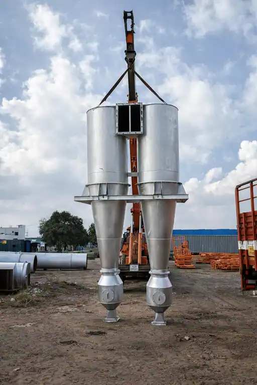 Twin high-efficiency silver cyclone dust separators suspended by a crane in an outdoor industrial fabrication yard